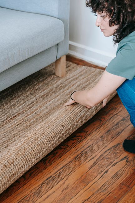 A woman with curly dark hair, dressed in a teal shirt and dark pants, is performing surface cleaning on a textured jute or natural fiber rug. She is kneeling on a polished wooden floor with a rich, reddish-brown tone and visible wood grains, using her hands to adjust or straighten the rug. The rug is positioned adjacent to a light gray upholstered sofa with wooden legs, in a well-lit room with neutral wall color. A spray bottle and cleaning cloth may be present but are not visible; the focus is on her careful handling of the rug. This scene illustrates a domestic cleaning process aimed at maintaining hygiene and appearance, consistent with deep cleaning and surface sanitisation services offered by Carpet Cleaners Colliers Wood, near Colliers Wood Station.