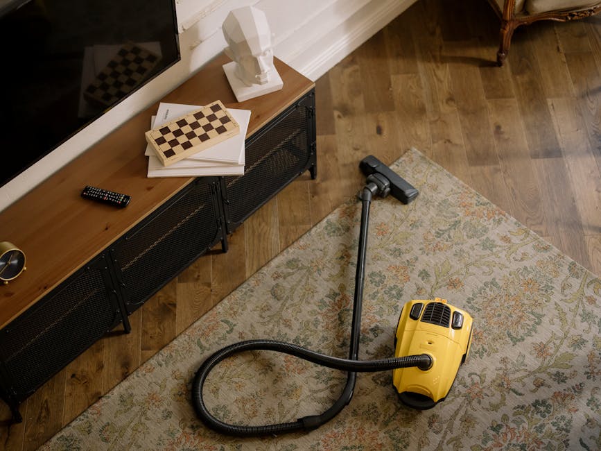 An indoor living room scene featuring a hardwood floor with a decorative patterned area rug. Next to the rug is a yellow handheld vacuum cleaner with its hose extended, indicating surface cleaning in progress. Against the wall, a black metal and wood media unit with a flat-screen television on top holds a white bust sculpture, a remote control, and some neatly stacked papers and books. The room is well-lit with natural light, highlighting the clean, polished appearance of the wooden floor and the tidy arrangement of items, reflecting professional domestic cleaning services by Carpet Cleaners Colliers Wood.