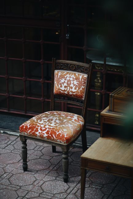 An antique-style chair with a wooden frame and ornate carved details, upholstered in cream and orange patterned fabric, is positioned on a tiled floor outdoors. Next to the chair is a wooden side table with a closed box on top, and behind it, a glass door with a grid pattern is visible, reflecting faint light. The scene appears clean and well-maintained, with natural lighting highlighting the textures and colors of the furniture. This setting suggests a focus on surface cleaning and upholstery care, consistent with services offered by Carpet Cleaners Colliers Wood, especially in a residential or commercial environment requiring deep cleaning or sanitisation of upholstered furniture.