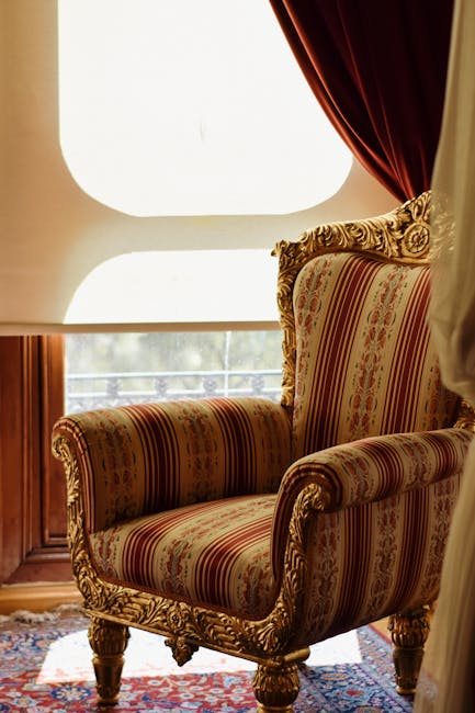 An ornate armchair with gold-accented carved wooden legs and a high, curved backrest upholstered in fabric with red and gold stripes and floral patterns. The armchair is positioned on a patterned area rug in front of a large, rounded window with white framing, allowing natural light to illuminate the room. To the right of the window, there are deep red velvet curtains, partially drawn to one side. The surrounding room and furniture suggest a well-maintained, elegant interior, consistent with domestic spaces serviced by Carpet Cleaners Colliers Wood for routine and deep cleaning needs.