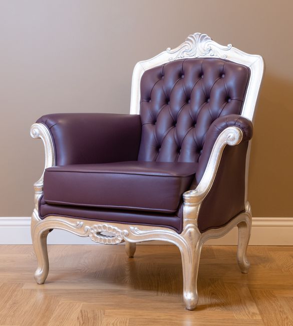 An antique-style chair with a wooden frame and ornate carved details, upholstered in cream and orange patterned fabric, is positioned on a tiled floor outdoors. Next to the chair is a wooden side table with a closed box on top, and behind it, a glass door with a grid pattern is visible, reflecting faint light. The scene appears clean and well-maintained, with natural lighting highlighting the textures and colors of the furniture. This setting suggests a focus on surface cleaning and upholstery care, consistent with services offered by Carpet Cleaners Colliers Wood, especially in a residential or commercial environment requiring deep cleaning or sanitisation of upholstered furniture.