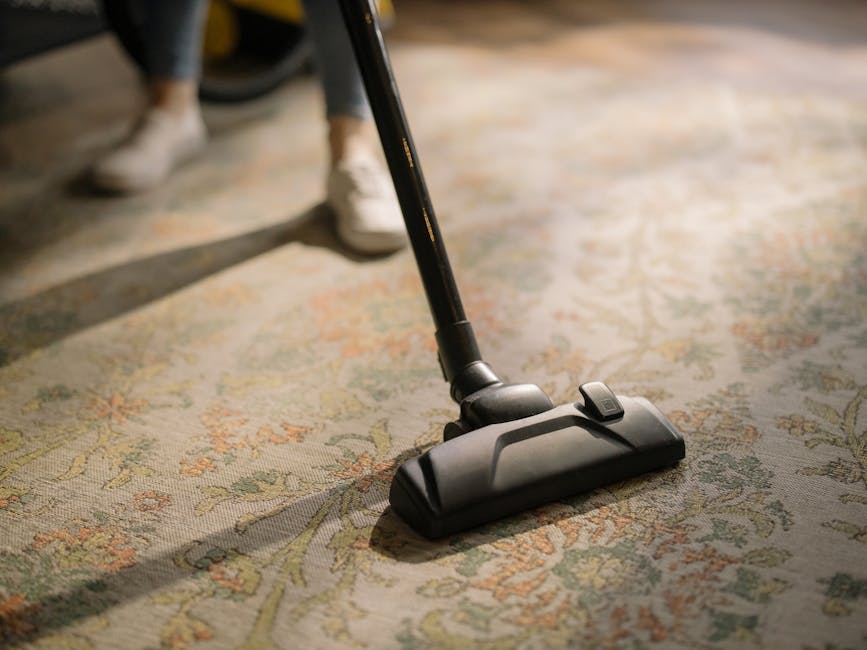 A person in black clothing is vacuuming a beige, textured area rug on a wooden floor using a red vacuum cleaner with a connected suction head. The vacuum cleaner features blue buttons and symbols indi